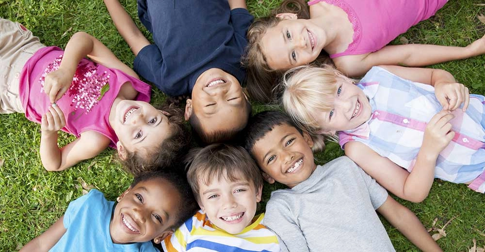 Seven smiling young children lay in circle, heads pointed toward each other, on the grass