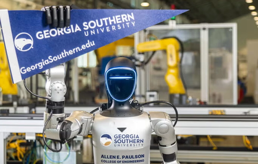 Photograph of a humanoid robot holding a Georgia Southern University pennant in a robotics lab. Robot features a sleek black face with blue LED lights, metallic arms, and a torso labeled with university and college of engineering logos, with industrial robotic arms visible in the background.