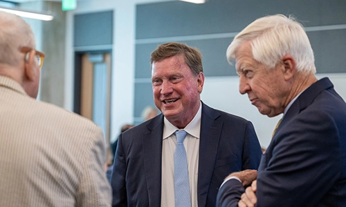 Photograph showing three older men in business attire conversing inside a modern office or conference venue. Center man smiles toward camera while two men, one partially visible at left and one with folded arms at right, engage in discussion.