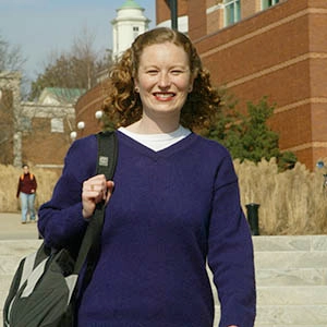 A student in a purple sweater wearing a book bag smiling at the camera as she walks towards it