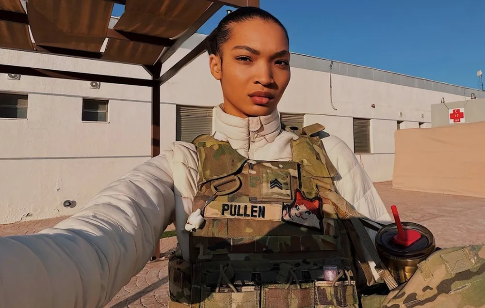 A woman in military gear takes a selfie outdoors in front of a white building. She is wearing a cream-colored puffer jacket under a camouflage tactical vest that features a name tape reading 