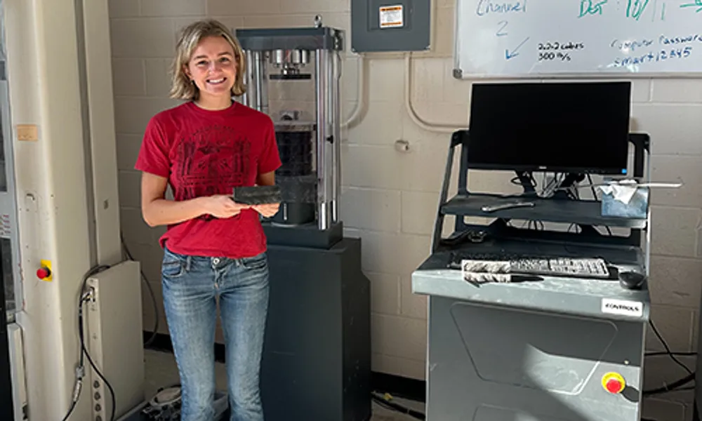 Photograph of a young woman standing in a lab setting, holding a rectangular object next to a tall cylindrical machine and a computer workstation. The scene highlights a research or testing environment with equipment likely used for material analysis or mechanical testing.