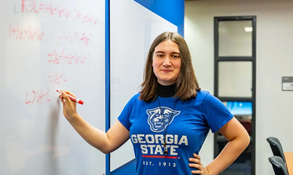 Photograph of a woman standing and writing on a whiteboard in a classroom or office setting. She wears a blue Georgia State University t-shirt and smiles at the camera, with mathematical or scientific equations written in red marker on the whiteboard.