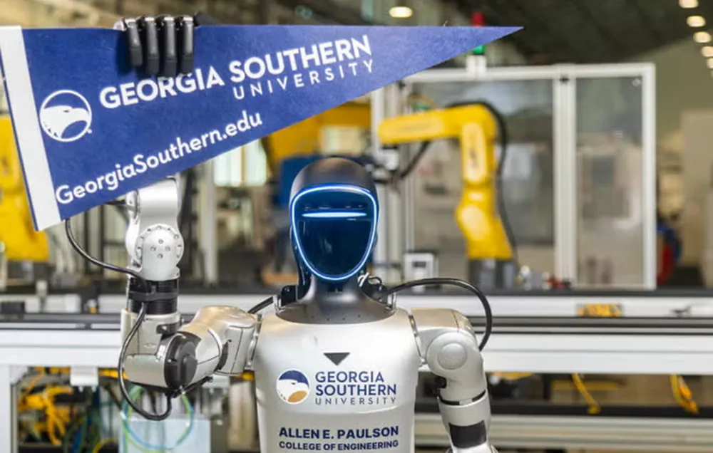 Photograph of a humanoid robot holding a Georgia Southern University pennant in a robotics lab. Robot features a sleek black face with blue LED lights, metallic arms, and a torso labeled with university and college of engineering logos, with industrial robotic arms visible in the background.