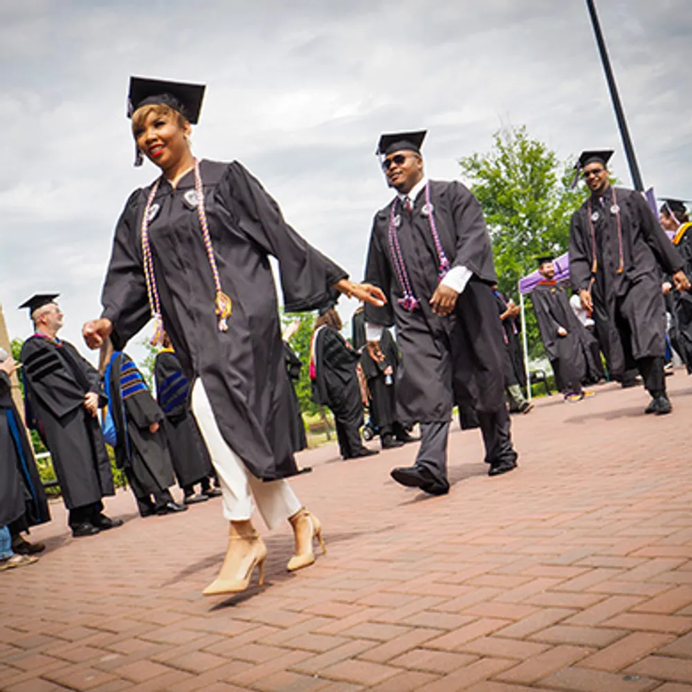 Students at graduation walking on a red brick pathway.  They are dressed in black graduation gowns.