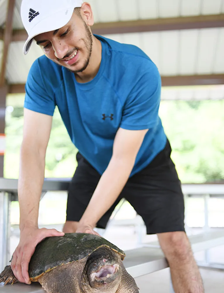 Student in a white cap and blue shirt smiling.  He's holding onto a turtle with both hards.