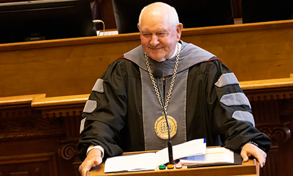 A person wearing academic regalia, including a black gown with gray velvet panels and a large medallion on a chain, standing behind a podium.