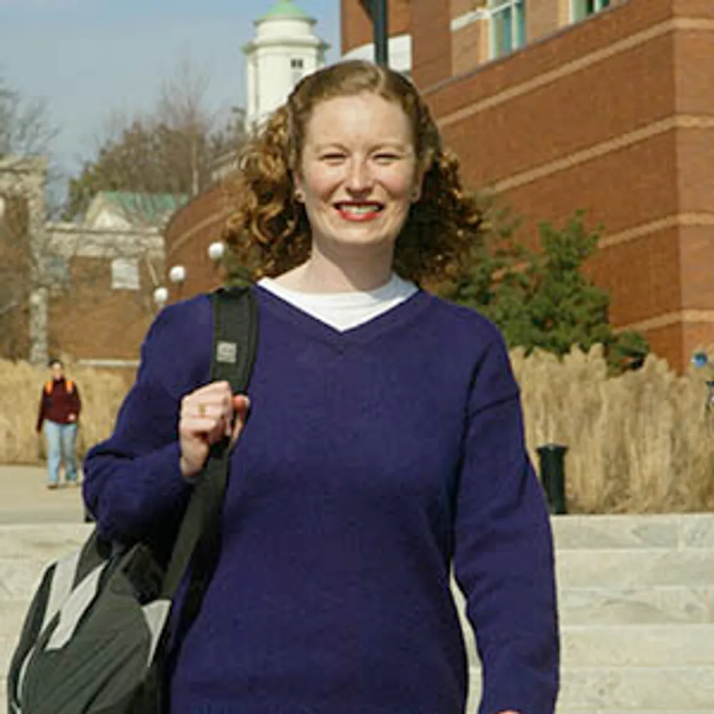A student in a purple sweater wearing a book bag smiling at the camera as she walks towards it
