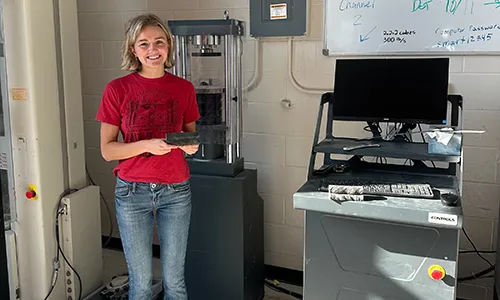 Photograph of a young woman standing in a lab setting, holding a rectangular object next to a tall cylindrical machine and a computer workstation. The scene highlights a research or testing environment with equipment likely used for material analysis or mechanical testing.