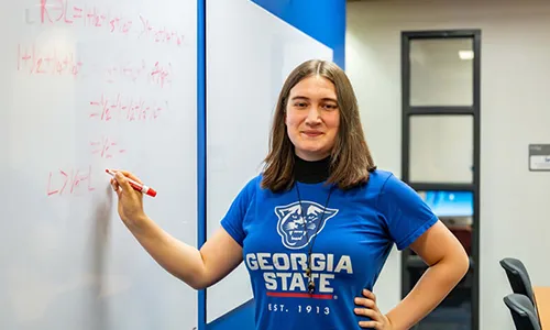 Photograph of a woman standing and writing on a whiteboard in a classroom or office setting. She wears a blue Georgia State University t-shirt and smiles at the camera, with mathematical or scientific equations written in red marker on the whiteboard.