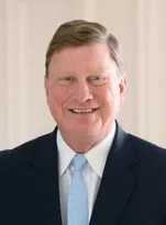 A smiling Tom Fanning in a dark pinstripe suit, white shirt, and light blue tie. Formal head-and-shoulders portrait presents a professional appearance against a neutral beige background.