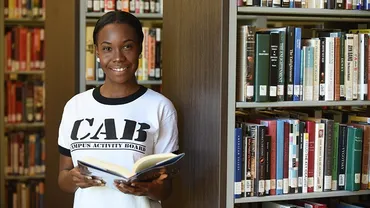 A female student in a white shirt holds an open book and leans against a bookshelf inside a library.