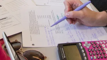 A hand holds a pen over a homework paper beside a calculator.