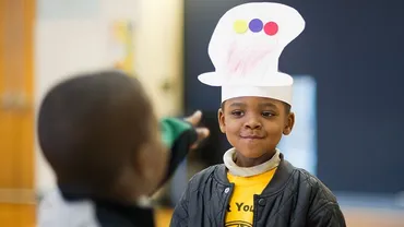 A child points at another child wearing a crafted hat made out of paper material.