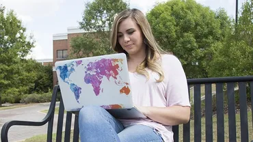 A woman sits on a bench outside, her legs are crossed and an open laptop on one knee. The back of the laptop has a colorful illustration of Earth's continents.