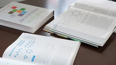 Three books lay on a wooden table, one closed and two open, with chemistry diagrams on their pages.