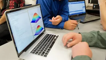 Two students sit at a table with two open laptops running various software.