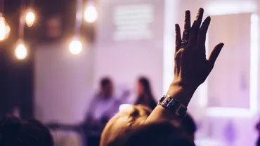 A person sitting as an audience member in a room holding their hand up to ask a question to the two presenters in the front of the room in front of them.